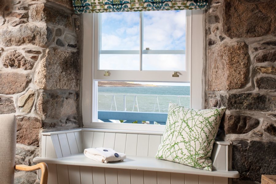 Cornish cottage windowseat with grainte stone walls either side, window open to show sea view, green and white cushion and folded blanket on the seat. Cornish Gems property "Trevanion Cottage" available for October half term.