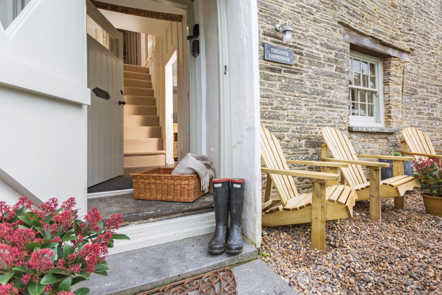 Cornish Gems property "Tregonce Farmhouse" available for October Half Term, photo of front door and porch with wellington boots and blanket in hamper on doorstep, door opened to show stairs. Three wooden chairs in front garden under the window in front of stone wall.