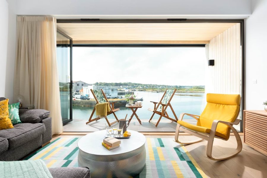 Living room with jellow chair, grey sofa and silver coffee table, wide open doors onto balcony overlooking waterfront of Hayle Cornwall, with two deck chairs and small table. Clearwater House let by Cornish Gems available for October half term.