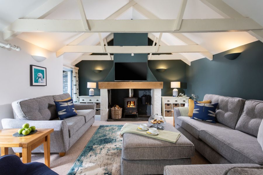 Cornish Gems property interiors of "The Linhay", a Cornish holiday home let by Cornish Gems for October half term. Photo shows living room with grey sofas, dark teal walls, lit log burner and tv over the mantlepiece. White wooden beams in the ceiling.