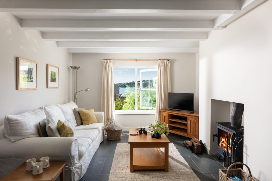 Cosy living room with sea view window, TV, sofa, coffee table and lit log burner at Cornish Gems property "Pandora Cottage" available for October half term