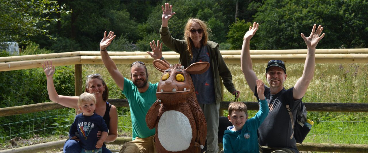 Photo from Forestry England of group of people waving their hands and smiling around a wooden statue of the character "Gruffalo's Child" by Julia Donaldson.