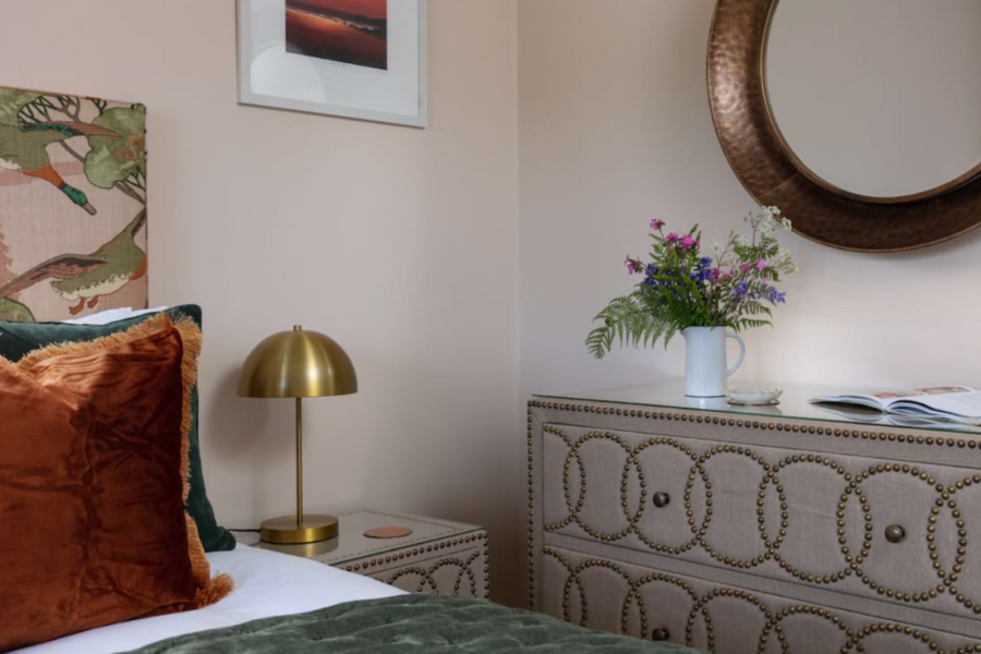 Cornish Gems property "Run Ashore" available for October half term image featuring interiors with orange and blue cushions on the bed, a gold bedside lamp, studded upholstered drawers, vase of wild flowers and a copper round mirror.
