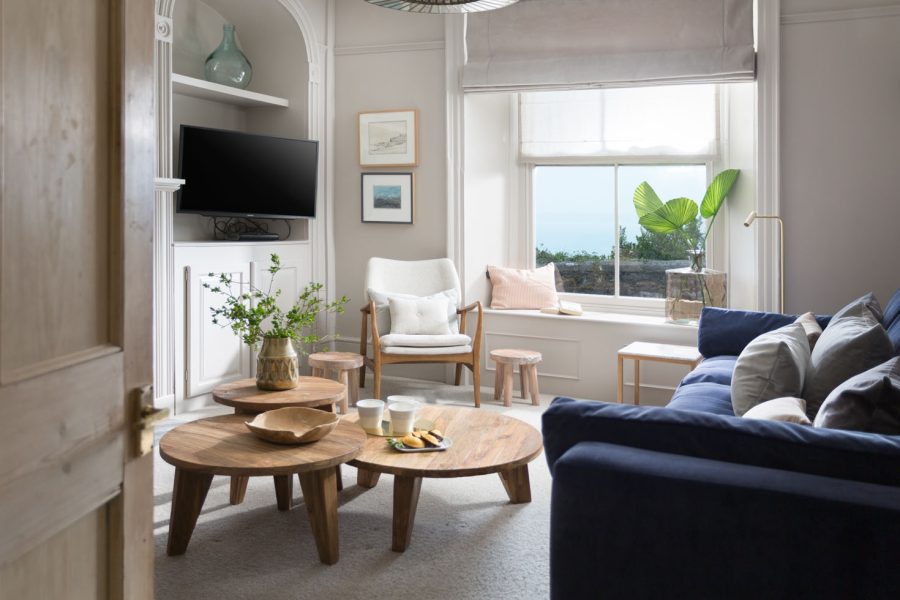 Living room of townhouse with three wooden coffee tables with mugs, bowl and vase of flowers. Navy blue sofa and plant on windowseat. with sea view. TV in alcove on wall. White chair by window. Property is Porthminster Townhouse in St Ives, Cornwall, let by Cornish Gems.