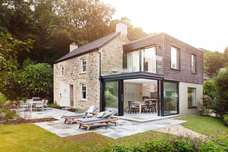 Extended Cornish cottage with large open bifold doors and stone cottage walls, sun loungers on patio and trees and lawn surrounding. Wren Cottage let by Cornish Gems set near St Agnes.