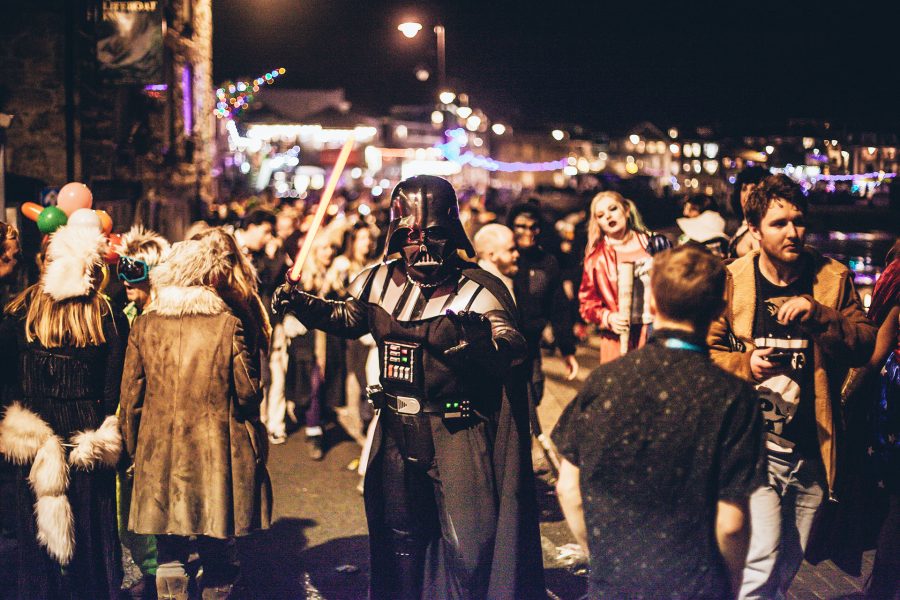 Fancy dress pub crawl, focus on punter dresses at Star Wars storm trooper, posing amongst crowd of others in fancy dress, on St Ives harbour front at night, lit up with Christmas lights