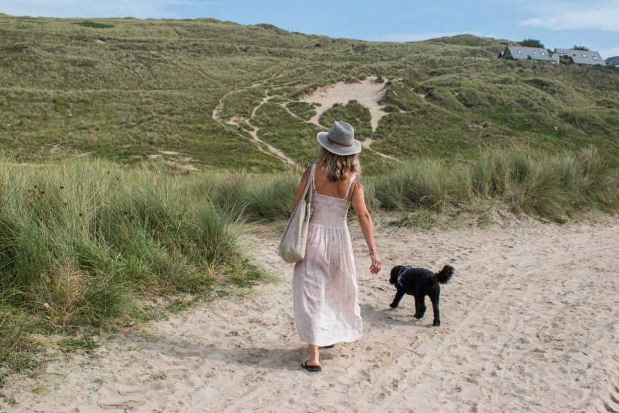 Woman in white dress and sunhat with tote bag walkng black dog on sand dunes in Cornwall on dog friendly walk on Cornish Gems holiday