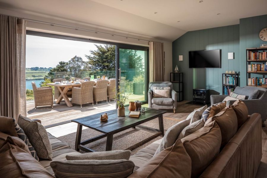 large living area with open trifold doors looking out to sea views, dining table on the balcony, coffee table and sofa to the freground, tv on blue wall to rear right 