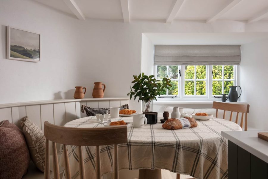 table made up with beige checkered table cloth, surrounded by bench build into wall, and wooden chairs. picture on frame on white walls, flowers and food on the table, window to the right, rear of the table, looking our to green hedge, with white ceiling and beams