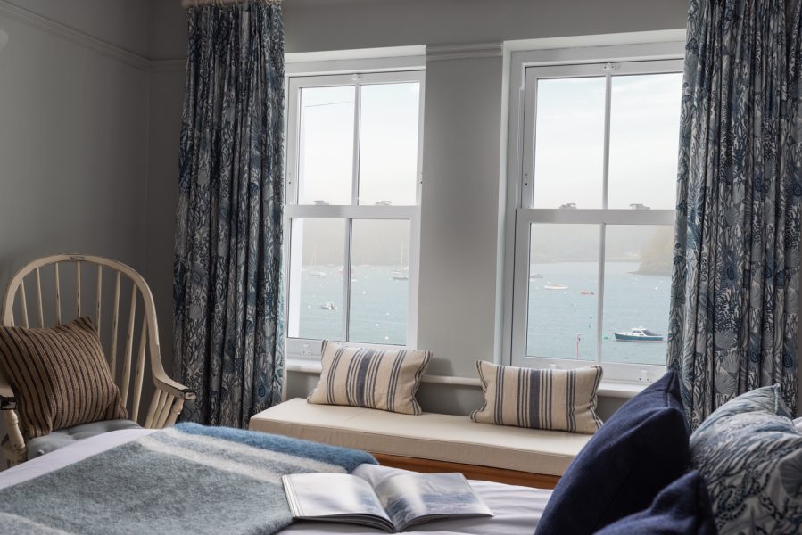 two pane windows with curtains drawn open to show moody, cloudy sea views over st mawes, window seat with cushions and armchair to the left. magazine open on bedspread in foreground, cushions to the right