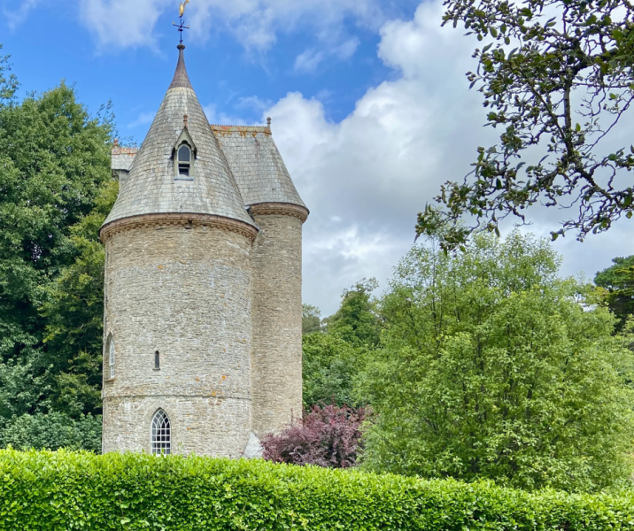 water tower like rapunzel with cone roof and rounded walls, to the left of the image, trees in back and foreground, hedge in front of building, blue sky with white clouds