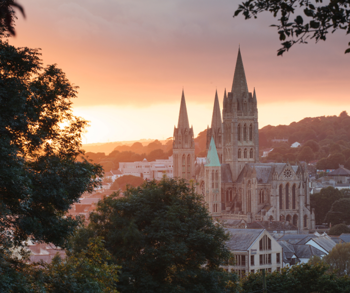 truro cathedral to the right, tree branches framing photo in foreground, all slightly silhouetted by sunrise over truro city